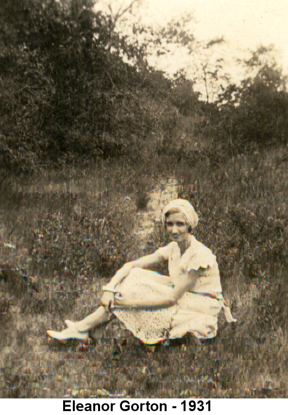 Black and white photo of Eleanor Gorton in a lacy white dress and white cap, bobbed hair peeking out, sitting with her knees up, arms wrapped around them, in a grassy field with trees in the background.